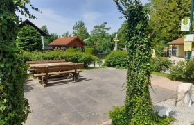 Frohsdorf, © Thermengemeinden A quiet square with wooden benches and tables, surrounded by green plants and an ivy-covered archway.
