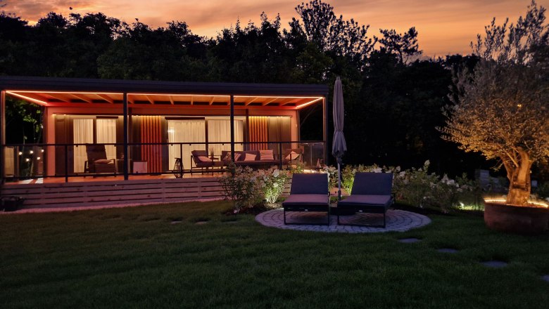 Johannesbachklamm Chalet, © Johannesbachklamm Chalet A modern chalet with terrace and sun loungers at sunset, surrounded by trees and an illuminated tree in the garden.