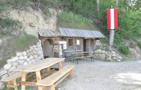 Cave, © ZVG Gemeinde A rustic wooden hut with a table and benches in front of it, built into a hill. A red and white flag hangs from a tree.