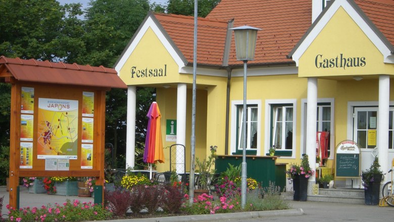 Municipality of Japons, © Gemeinde Japons Yellow building with the inscription 'Festsaal' and 'Gasthaus', surrounded by flowers and an information sign.
