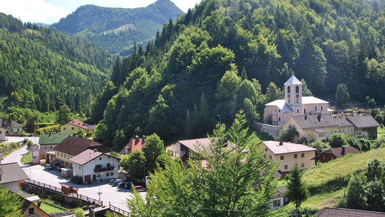 Location photo, © Gemeinde Schwarzenbach/Piel. Small village in a green mountain landscape with a church and houses.