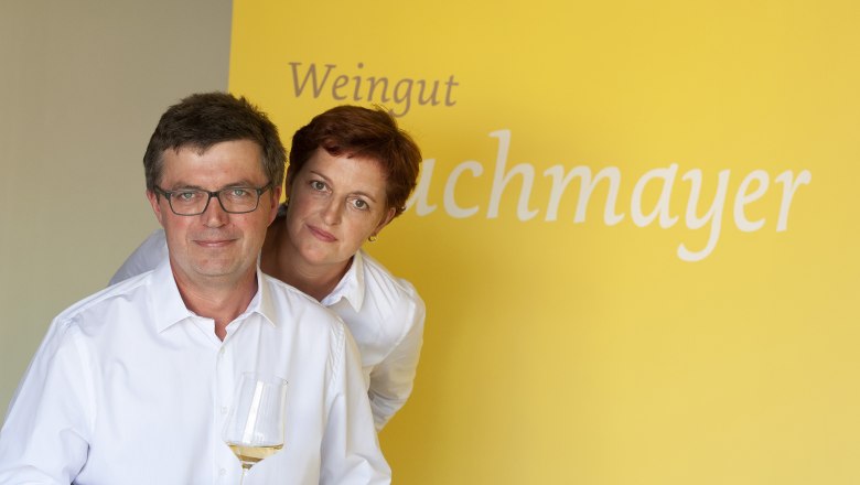 Maria and Thomas Buchmayer, © Michael Himml Two people in white shirts in front of a yellow background with the inscription 'Weingut Buchmayer'. One person is holding a wine glass.