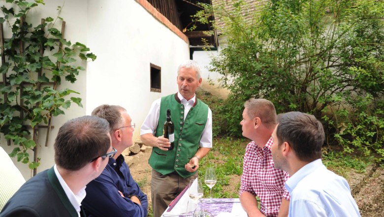 Guests at the Honisg-Keller, © H. Schleich A man in traditional dress presents a bottle of wine to a group of four men at an outdoor table.