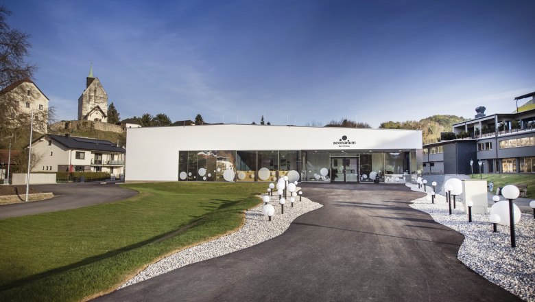Event center with interactive exhibition "Sconarium", © Gemeinde Bad Schönau, Stefan Knittl Modern Sconarium building with glass front and decorative globe lights, surrounded by green spaces and the fortified church in the background.