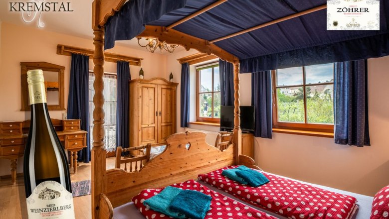Room no. 3 four-poster bed, © Weingut Zöhrer A cozy room with a four-poster bed, wooden furniture and a view of the vineyards. A bottle of wine is pictured in the foreground.