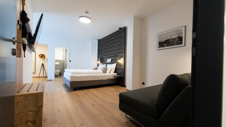 VinoQ, © ohida OG / ohne.media Modernly furnished hotel room with double bed, sofa and flat-screen TV. Light wooden floors and a bathroom in the back.