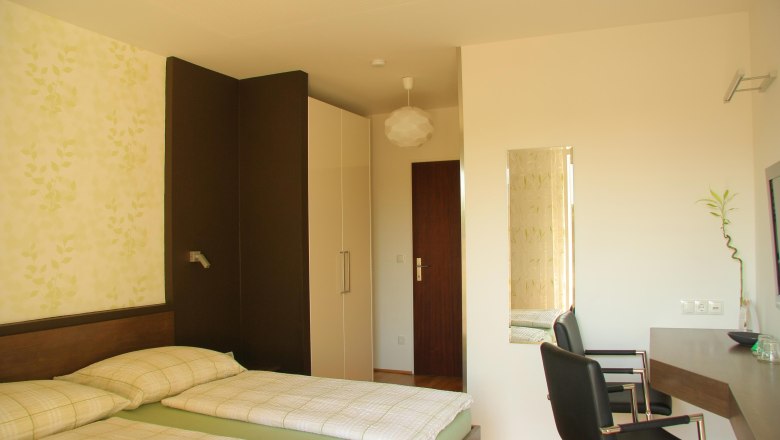 Comfort room, © Susanne Kernbichler A modern hotel room with two single beds, a desk and a chair. The walls are bright and decorated with a plant pattern.