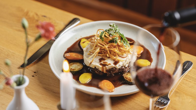 Ochsenschlepp in Zweigeltsauce with mashed potatoes, © Niederösterreich Werbung/Michael Reidinger A plate of oxtail in two-yolk sauce, garnished with mashed potatoes and vegetables, on a wooden table. A glass of red wine is poured.