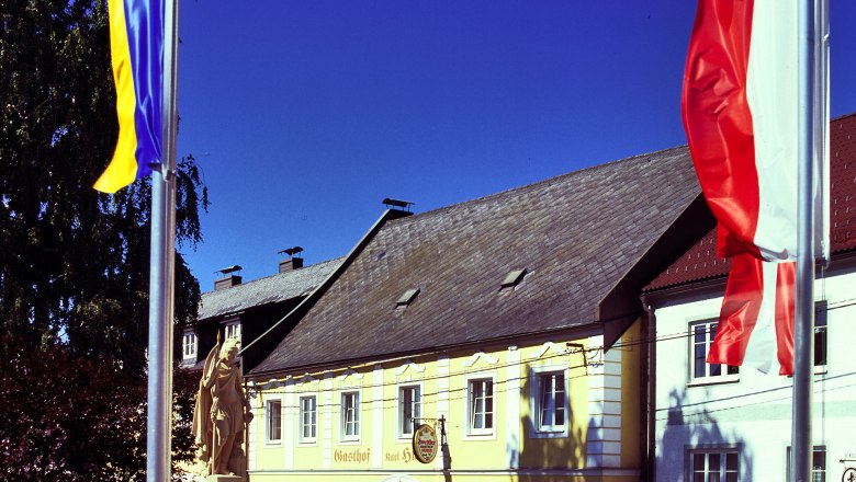 Gasthof Huber, © MG Rfd Yellow building with the inscription 'Gasthof Karl Huber', surrounded by flags and a statue.
