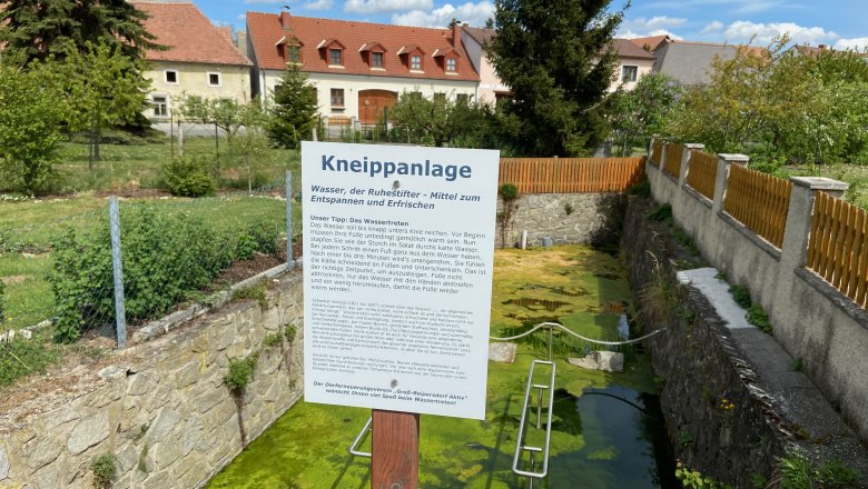 Water treading facility Großreipersdorf, © Weinstraße Weinviertel Kneipp facility with information board and water basin, surrounded by a fence and houses in the background.
