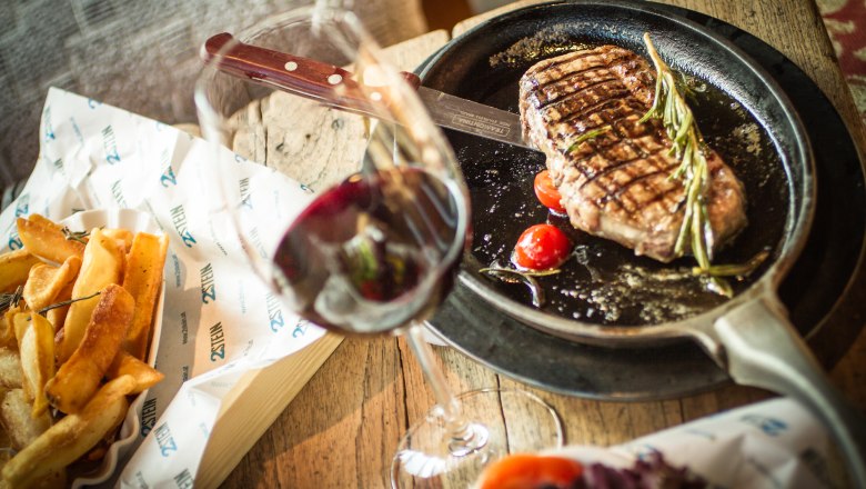 2Stein Bar-Café-Restaurant, © 2stein_Kastner new media A steak in a pan with tomatoes and rosemary, French fries and a glass of red wine on a wooden table next to it.