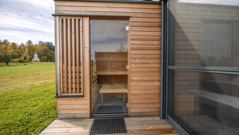 roo'n, © Niederösterreich Werbung / Maximilian Pawlikowsky Wooden building with glass door and view of a meadow and church in the background.