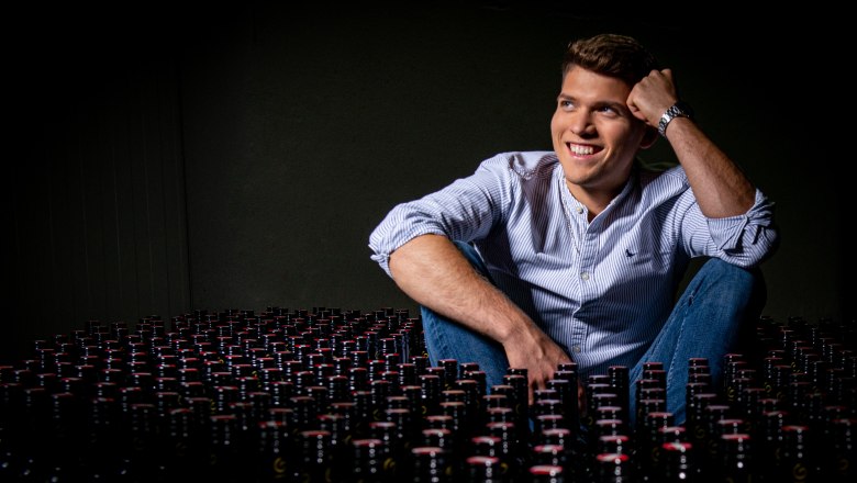David Gutmayer, © Martin Lifka Photography A man sits smiling in a wine cellar, surrounded by many bottles of wine.
