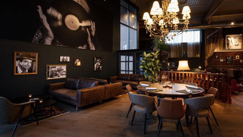 Zündwerk Living Room, © DERENKO DESIGN A stylishly furnished room with leather sofas, a round table and a large chandelier. Black and white photographs hang on the wall.
