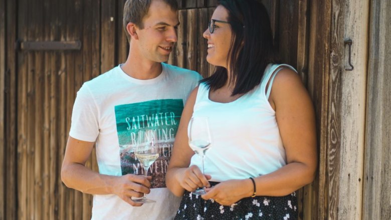 Markus Heninger and Lisa, © Furtmüller Fotografie A man and a woman stand smiling in front of a wooden wall, holding glasses of wine.