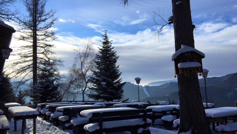 Terrace Speckbacher Huts, © Karin Stranz Snow-covered terrace with wooden benches and tables, surrounded by trees and mountains in the background.