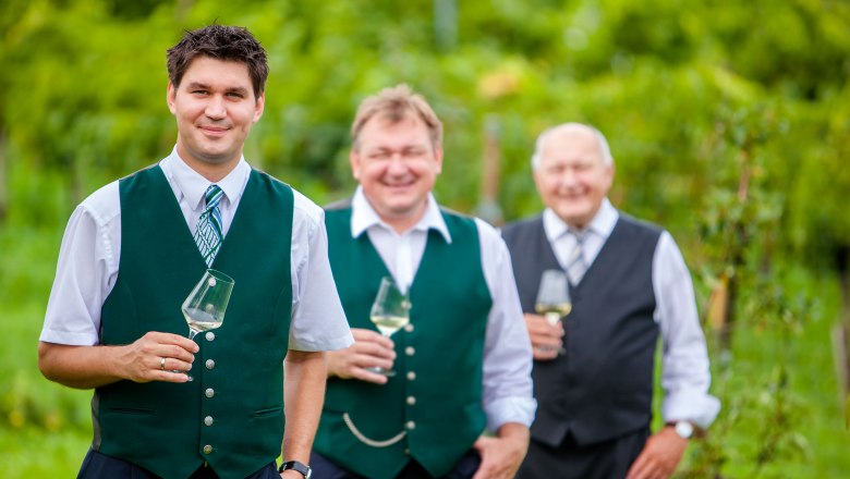 Müllner family, © Michael Müllner Three men in green vests hold glasses of wine in a garden.