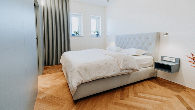 Newly adapted rooms invite you, © Der Löffler - Nicole Seiser Modern bedroom with double bed, parquet floor and gray curtains.