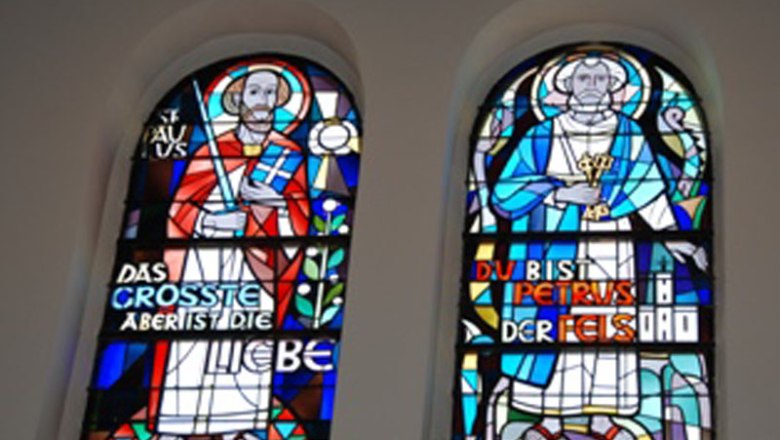 Windows, © Pfarre St. Jakobus Two colorful stained glass windows with religious figures and inscriptions.
