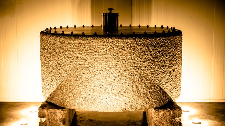 Pressing stone, © Bernhard Kastner A large, cylindrical pressed stone made of coarse material, illuminated by warm light, stands on two stones in a room.
