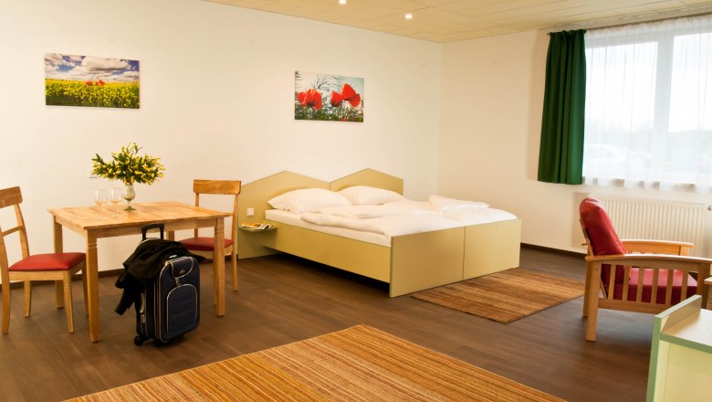 Studio, © Winrooms Betriebs GmbH A modern studio with double bed, table and chairs, red armchair and suitcase. Light-colored walls, green curtains and flower pictures on the wall.