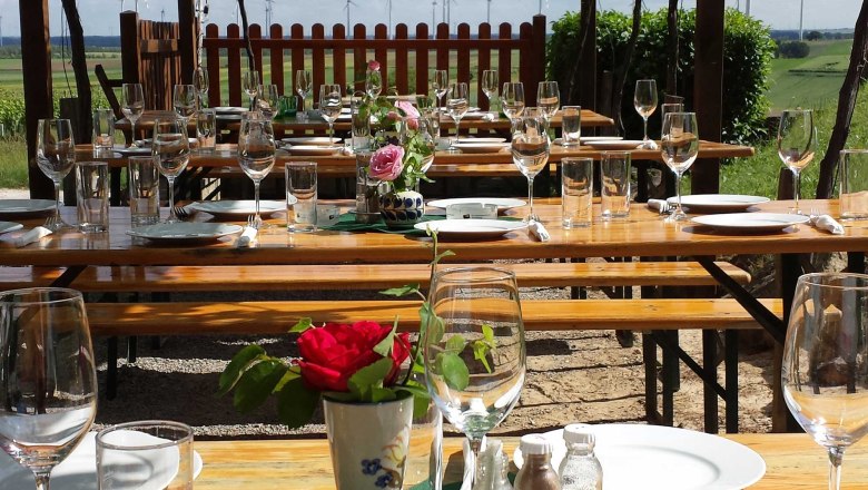 Buschenschankt Gratzer-Sandriester, © Weinbau Gratzer-Sandriester Tables set outside under a leafy canopy with a view of wind turbines in the distance.