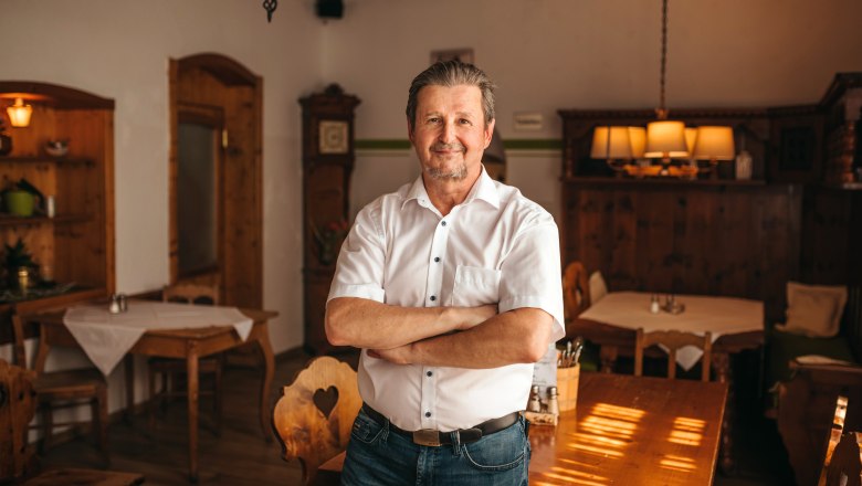 Landlord Florian Matschinegg, © Niederösterreich Werbung/Daniela Führer A man in a traditional dining room with wooden furniture, dressed in a white shirt and jeans, stands with his arms folded and smiles.