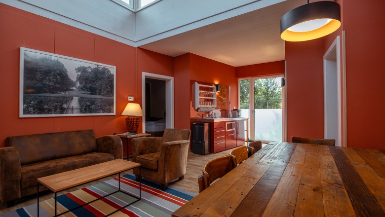 Grafenegg Cottages, © Niederösterreich Werbung / Maximilian Pawlikowsky Interior view of a modern living room with red walls, wooden table, sofa and kitchenette.