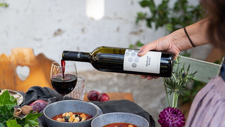 Wine enjoyment, © Carletto Ferrari A hand pours red wine into a glass next to two bowls of soup on a table.