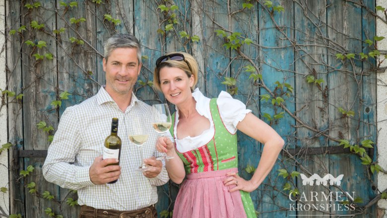 Winegrower couple, © Carmen Kronspiess A man and a woman in traditional dress hold wine bottles and glasses in front of an overgrown wooden wall.