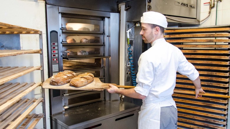 Bakehouse at Cafe Lechner, © Bäckerei Lechner A baker in white clothes puts bread into an oven.