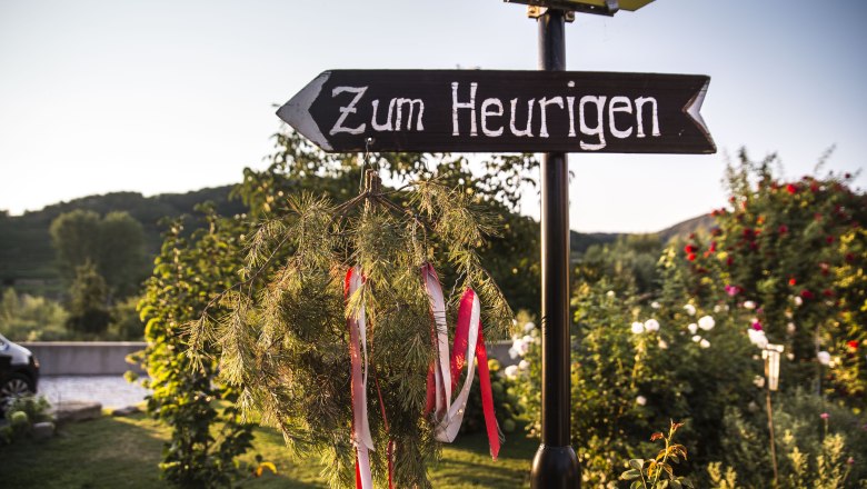 Signpost to the wine tavern Polz, © Daniela Matejschek A signpost with the inscription 'Zum Heurigen' points in one direction. Trees and flowers can be seen in the background.