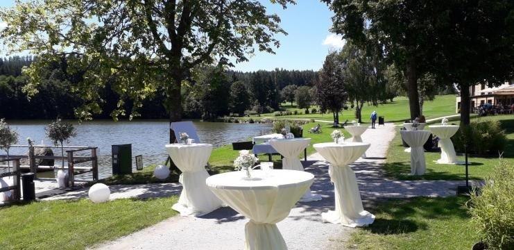 Hotel-Restaurant Hausschachen, © Hotel-Restaurant Hausschachen Bar tables with white cloths on the shore of a lake, surrounded by trees.