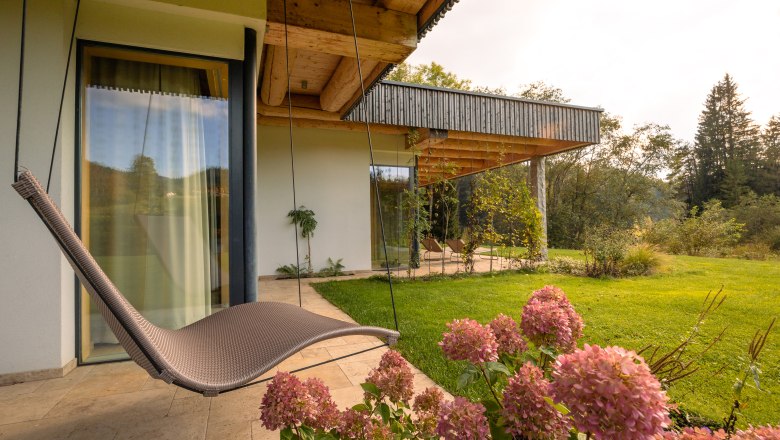 Vacation home by the lake, © Niederösterreich Werbung / Maximilian Pawlikowsky A modern vacation home with wooden veranda, hanging chair and blooming hydrangeas in the foreground, surrounded by green nature.