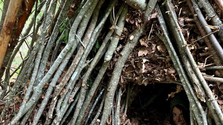 Survival Workshop Wilderness Center Nasswald, © Georg Bergthaler A person peers out of a self-made shelter made of branches and leaves in the forest.