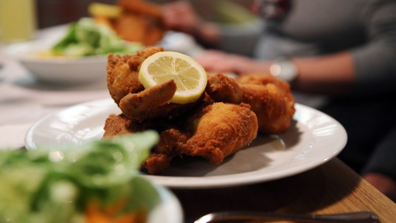 The famous fried chicken at Waldgasthof Steinbründl, © Familie Kogler-Renner A plate with breaded fried chicken and a slice of lemon, with a person and salad in the background.