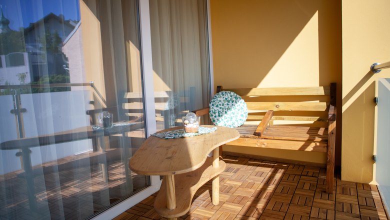 Balcony, © Michael Sokolar A balcony with a wooden floor, a wooden table and a wooden bench with a round cushion. A glass railing and a building can be seen in the background.