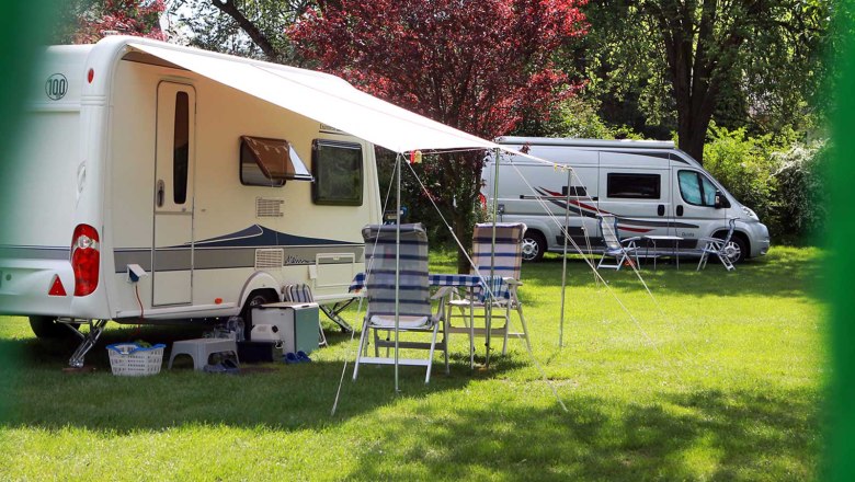 Campsite, © Szilágyi Caravan and motorhome on a green campsite with chairs and table under an awning.