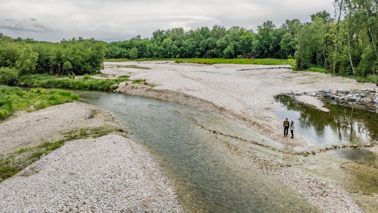 At the source of the Leitha in Lanzenkirchen, © Wiener Alpen/Fülöp, Kremsl A wide gravel bed of a river with clear water flowing through it. Two people standing by the river, green forest in the background