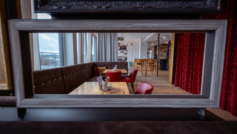 The Alfred, © Niederösterreich Werbung / Maximilian Pawlikowsky Interior view of a stylish café with red chairs and wooden tables, seen through a picture frame.