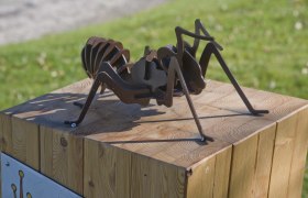 Corona's ant trail, © Martin Weiner Metal sculpture of an ant on a wooden block outdoors.