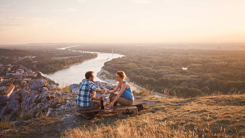 View from Braunsberg, Hainburg/Danube, © Donau Niederösterreich, Andreas Hofer Two people sitting on a hill overlooking a river and a city at sunset.