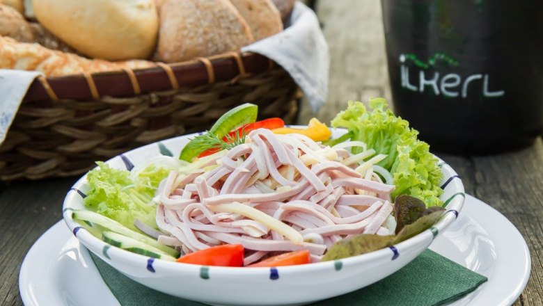 Sausage salad, © Weinberghof Ilkerl A plate of sausage salad, garnished with lettuce leaves, tomatoes and cucumber. A bread basket in the background.
