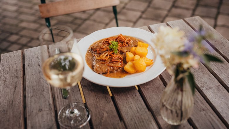 Stewed roast onion of Angus beef with roast potatoes, © Niederösterreich Werbung/Daniela Führer A plate of roast onions and roast potatoes on a wooden table, next to it a glass of white wine and a vase of flowers.