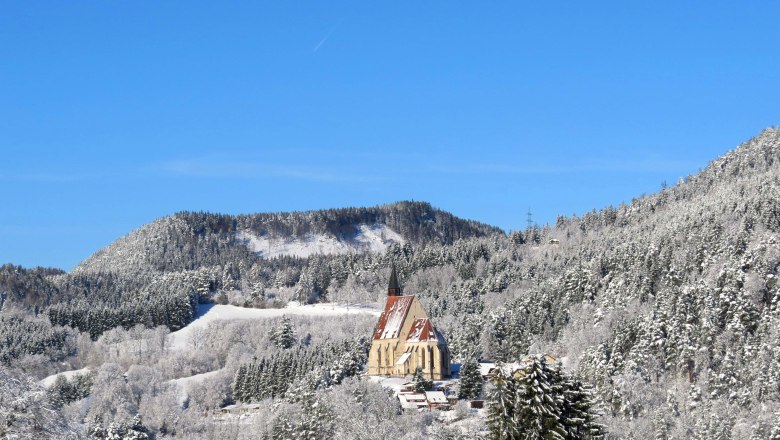 St. Wolfgang's Church towers over the village, © Marktgemeinde Kirchberg, Wolfgang Riegler Winter landscape with a church in snow-covered mountains under a blue sky.