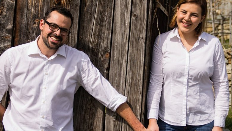 Barbara and Erwin Tauchner, © Weingut Tauchner Two people in white shirts stand in front of a wooden wall and smile.