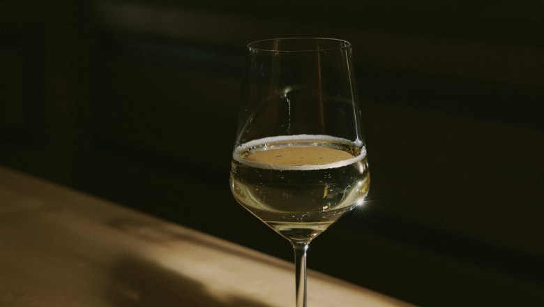 Wine glass, © Weinviertel Tourismus / Sophie Menegaldo A wine glass with white wine stands on a wooden table, illuminated by a warm beam of light.