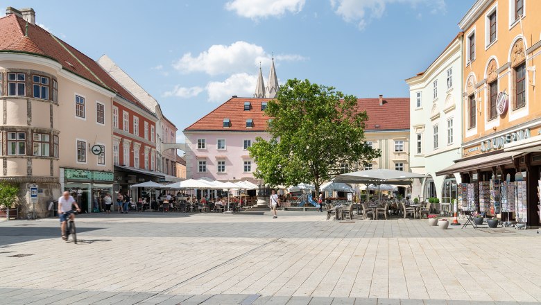 Main square with Café Ferstl, © Wiener Alpen/Christoph Schubert A lively main square with cafés and stores, surrounded by historic buildings. People sit outside, a cyclist rides past. Blue sky with clouds.