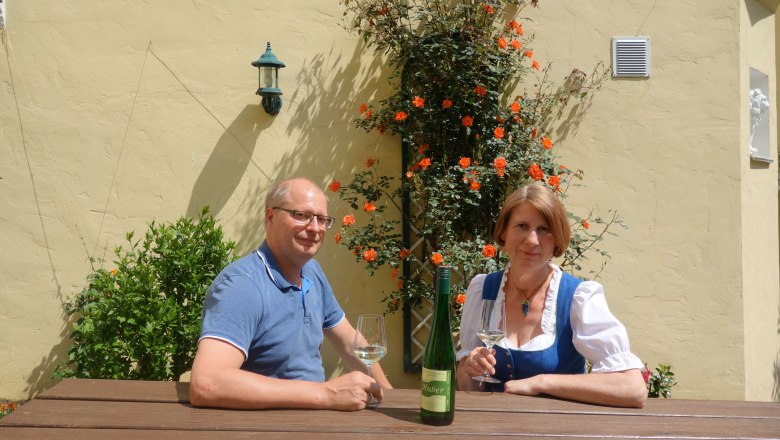 Operator couple Huber, © Familie Huber A man and a woman are sitting at a wooden table outside, with wine glasses and a bottle of wine. Orange flowers bloom in the background.