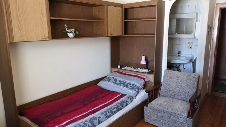 Single bed, © Gudrun Rath Single bed with red and gray bed linen, an armchair next to it and a washbasin in the background.